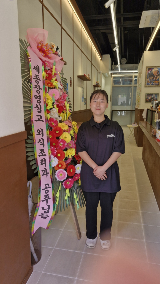 신나혜 사장이 장영실고 후배들이 보내준 개업기념 축하화환 옆에서 기념촬영을 하고 있다. 사진=강대묵 기자