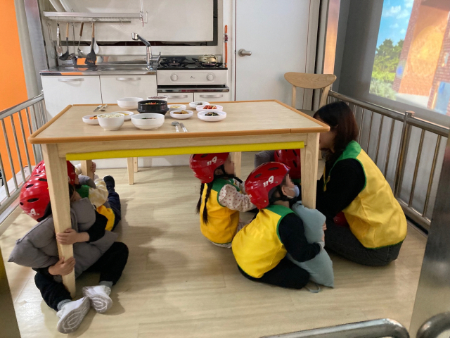 ▲아이들이 지진 발생 상황을 체험하며 책상 아래로 몸을 숨기고 머리를 보호하는 훈련을 하고 있다.